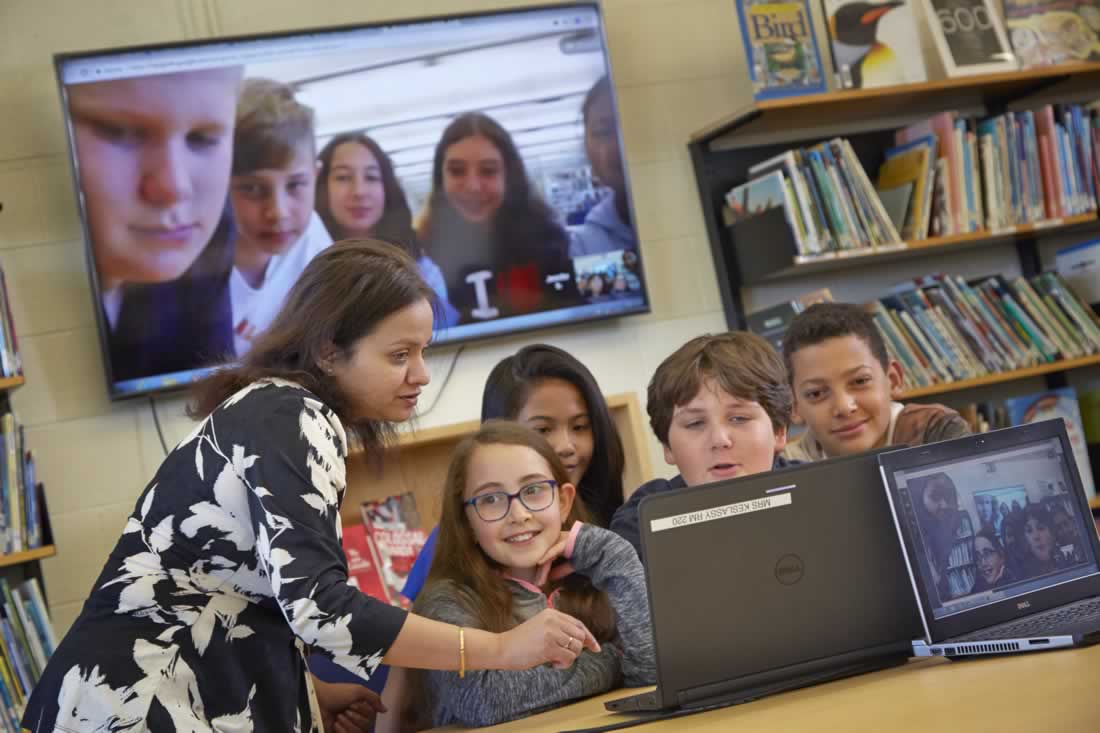 This is an image of a teacher helping a group of students working on a laptop with another group projected on a large tv on in the background'.